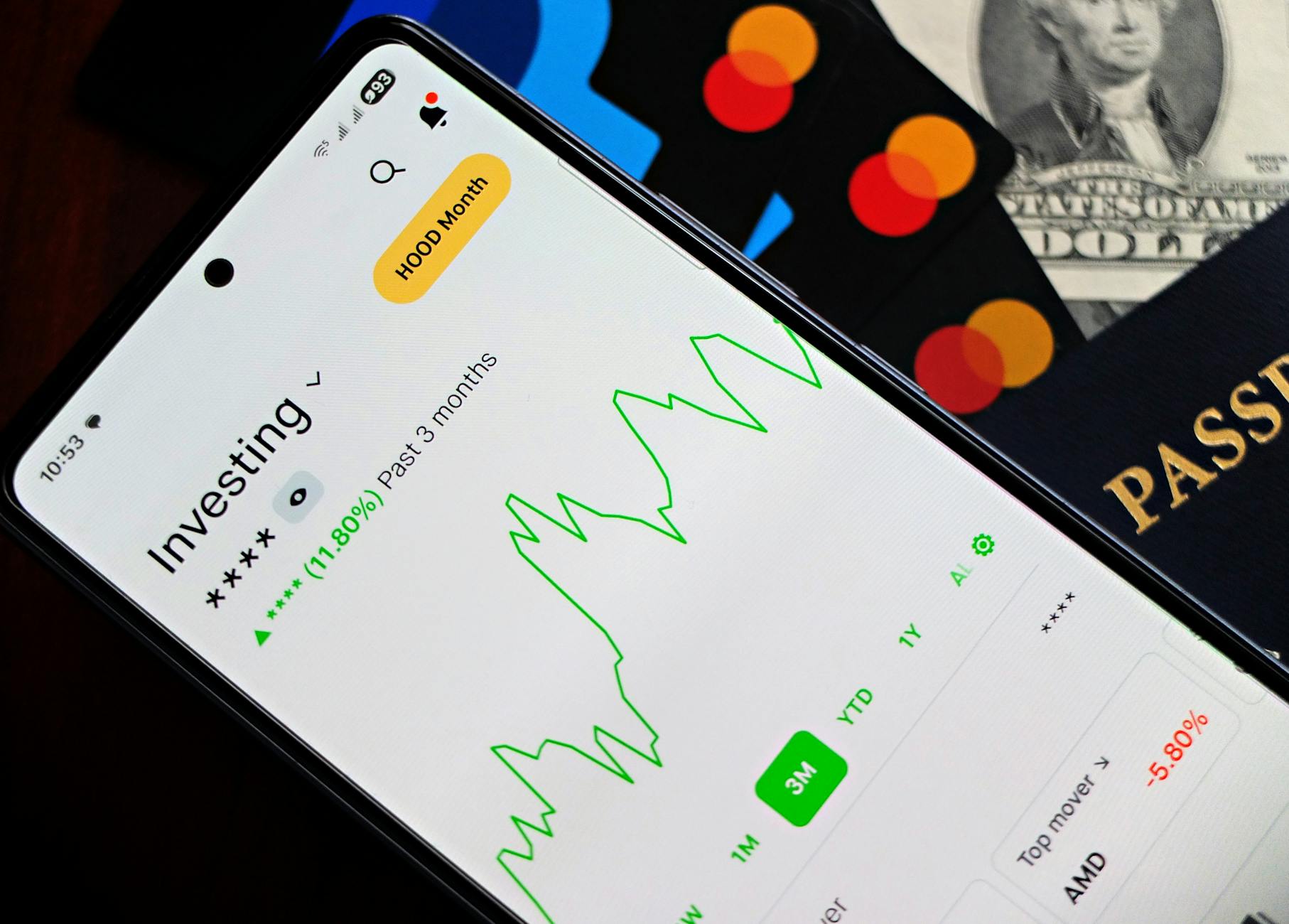 A smartphone showing an investment app with green growth indicators, surrounded by credit cards, US dollars, and a passport.